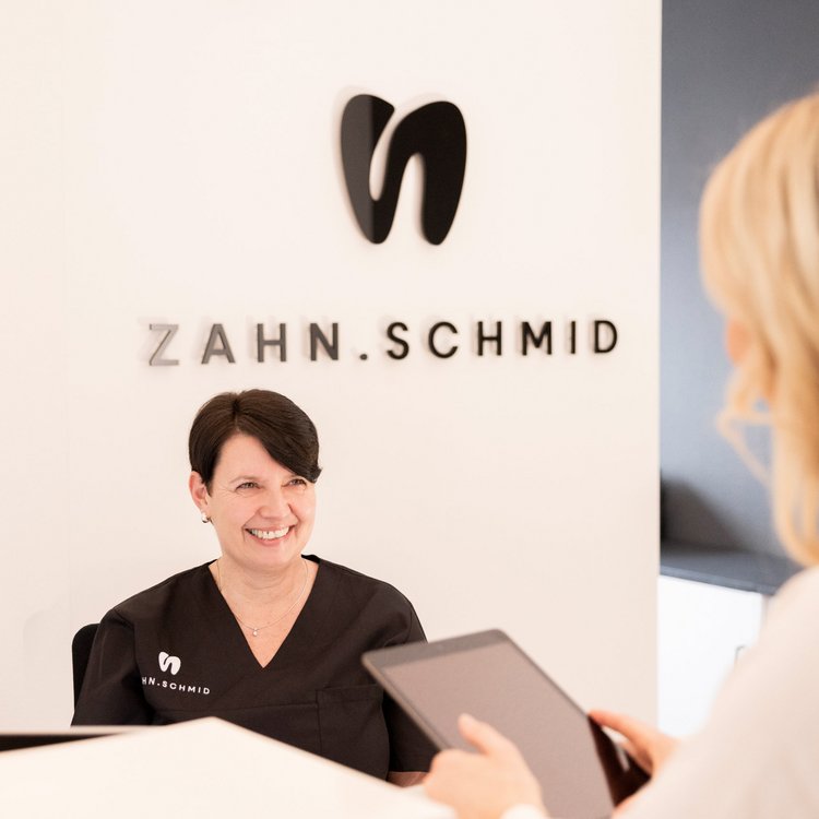 Herzlicher Empfang bei ZAHN.SCHMID Freundliche Empfangsmitarbeiterin der Zahnarztpraxis ZAHN.SCHMID begrĂŒĂt Patientin am Empfangstresen.